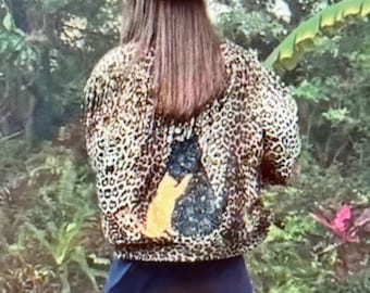 May include: A tan straw hat with a woven crown and a leopard print jacket featuring a large graphic of a cat on the back. The jacket is paired with a solid dark blue skirt. The ensemble is captured from behind, showcasing the animal print pattern.