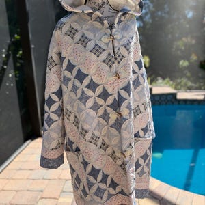May include: A hooded coat crafted from quilted fabric featuring a blue and white pattern of interlocking circles and plaid squares. The garment includes three wooden toggle closures down the front and two side pockets, with a light pink floral trim.