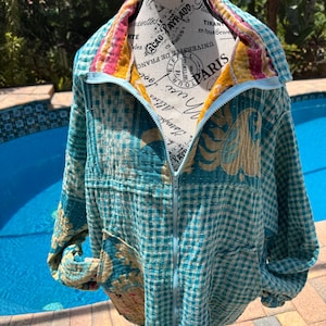 May include: A long-sleeve zip-up jacket featuring a quilted texture with a light blue and white gingham pattern. The garment includes a high collar, a central zipper, and visible stitching throughout, with a yellow and pink interior lining.