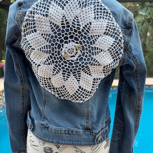 May include: A blue denim jacket featuring a large, circular white crocheted doily appliqued onto the back panel. The doily has an intricate, openwork floral pattern with two small, decorative gold-toned embellishments at the center.
