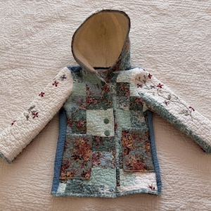 May include: A quilted hooded jacket featuring a patchwork design in shades of blue, green, and cream. The body displays a floral pattern, while the long sleeves are white with embroidered red flowers. It includes two front pockets and buttons.