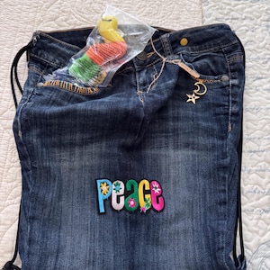 Op de afbeelding: Een denim trekkoordtas gemaakt van jeans, met een kleurrijke "peace" patch. De tas heeft een goudkleurige bedel met een ster en een maan. Een klein, verpakt speeltje is zichtbaar in de tas.