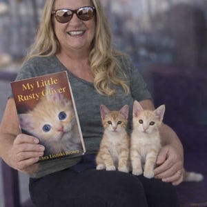 May include: A woman holding a book titled "My Little Rusty Oliver" with a kitten on the cover. Two orange tabby kittens sit on her lap. The book cover features a close-up of a kitten with blue eyes.