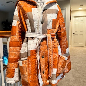 May include: A hooded jacket constructed from a patchwork of quilted fabric in shades of burnt orange, white, and tan. The garment features a tie-waist belt, two front patch pockets, and three dark metallic buttons down the center front closure.