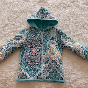 May include: A quilted jacket with a hood, featuring a teal, white, and coral paisley pattern. The jacket has a teal lining in the hood and a full-length zipper. The cuffs and trim are teal, and the jacket is laid flat on a beige surface.