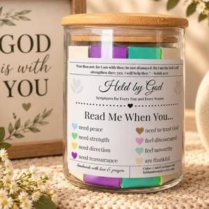 May include: A clear glass jar with a bamboo lid containing colourful paper slips. The label reads: Held by God, Scriptures for Every Day and Every Season, Read Me When You... need peace, strength, direction, reassurance, trust God, feel discouraged, feel unworthy, or are thankful.