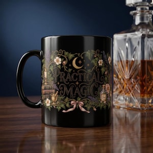May include: A black ceramic mug featuring the text "PRACTICAL MAGIC" in gold lettering. The design includes illustrations of a crescent moon, stars, a stack of books, a mortar and pestle, a small bottle, flowers, vines, and a pink ribbon.