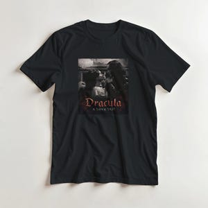 Könnte beinhalten: Schwarzes T-Shirt mit einem Graustufenbild von zwei sich umarmenden Personen, mit dem Wort "Dracula" in roter Schrift über "A LOVE TALE". Das Bild ist auf dem T-Shirt zentriert.