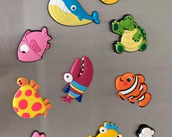 May include: A collection of ten colorful rubber sea creature magnets including a yellow fish, a blue whale, a pink fish, a green turtle, a yellow and pink spotted fish, a striped shark, an orange clownfish, a pink crab, a yellow seahorse, and a blue starfish.