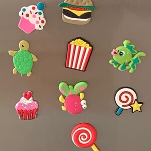 May include: A collection of colorful, cartoon-style magnets. The magnets depict a cupcake, burger, turtle, popcorn, fish, another cupcake, a bug, a lollipop, and a swirl lollipop.