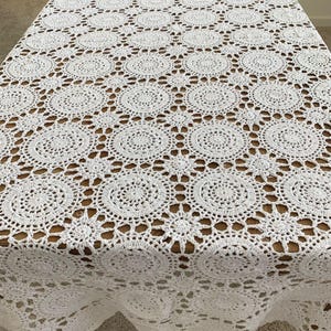 May include: A white, crocheted tablecloth with a repeating circular pattern. The intricate design features openwork circles and star-shaped accents, creating a delicate and textured surface. The tablecloth is draped over a wooden table.