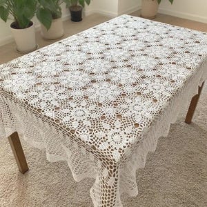 May include: A white, rectangular crochet tablecloth with a floral pattern covers a wooden table. The tablecloth has a scalloped edge and is draped over the table, showcasing the intricate openwork design. The table is set on a light-colored carpet.