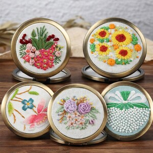 May include: Five round, bronze-framed compact mirrors with embroidered floral designs. Each mirror features a different colorful flower arrangement, including sunflowers, roses, and other blooms, set against a white or light blue background.