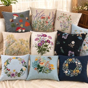 May include: A collection of nine square throw pillows featuring various embroidered botanical designs. The pillows display patterns of mushrooms, lavender, wildflowers, daisies, and floral wreaths in colors including gray, beige, black, and light blue.