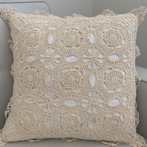 May include: A square throw pillow featuring a cream-colored crocheted cover with an intricate floral and geometric pattern. The delicate needlework is layered over a solid white fabric base, creating a textured appearance with scalloped edges.