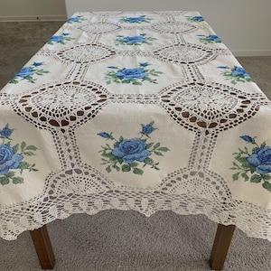May include: A rectangular tablecloth featuring an off-white fabric base with a repeating pattern of blue rose cross-stitch embroidery. The design includes intricate white crocheted lace panels between the floral sections and a scalloped lace border.