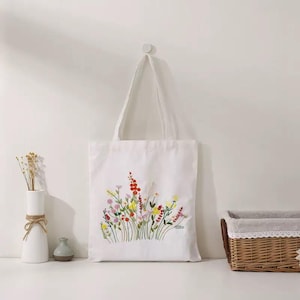 May include: White tote bag with an embroidered floral design in red, yellow, pink, and green. The bag has a simple, rectangular shape with long handles. The embroidery depicts a cluster of wildflowers.