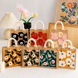 May include: A collection of seven small jute tote bags with two top handles, each featuring distinct tufted floral patterns in various colors including black, red, pink, yellow, dark blue, and white, arranged on a white surface.