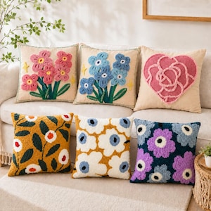 May include: Six square throw pillows with tufted textures. Top row: pink flowers, blue flowers, and a pink heart on beige fabric. Bottom row: mustard yellow with dark leaves, white with blue flowers, and dark blue with purple flowers.