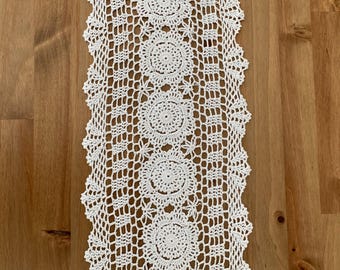 Può includere: Runner da tavolo bianco all'uncinetto con un motivo floreale ripetuto. Il runner è rettangolare e presenta un delicato design traforato. I bordi sono smerlati, aggiungendo un tocco decorativo. Il runner è esposto su una superficie di legno.