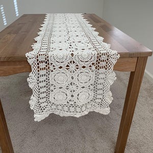 May include: A cream-colored, crocheted table runner with a detailed floral and geometric pattern. The runner is placed on a wooden table, showcasing its intricate design and texture. The runner's edges have a scalloped trim.
