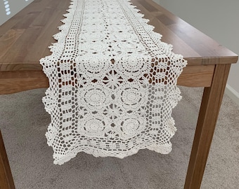 Può includere: Runner da tavolo all'uncinetto color crema con un motivo floreale e geometrico dettagliato. Il runner è posizionato su un tavolo di legno, mettendo in risalto il suo intricato design traforato e i bordi smerlati. Aggiunge eleganza a qualsiasi sala da pranzo.