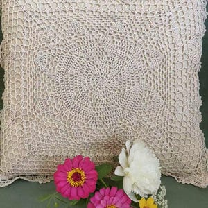 May include: A square, cream-colored crochet pillow with a detailed circular pattern. The pillow is adorned with a border of delicate crochet work. Bright pink and yellow flowers with green leaves are arranged in front of the pillow, creating a vibrant contrast.