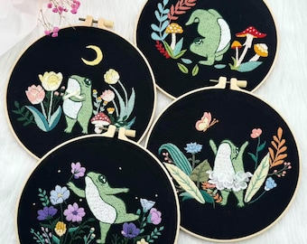 May include: Four round embroidery hoops with black fabric featuring whimsical frog designs. Each hoop showcases a different frog scene with colorful floral and mushroom accents. The frogs are depicted in various poses, adding a playful touch to the artwork.
