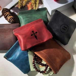 Puede incluir: Una colección de bolsas de cuero en varios colores, incluyendo marrón, rojo, verde, azul y negro. La bolsa roja presenta un diseño de cruz negra. Una bolsa está abierta, revelando un collar de cuentas y billetes doblados.