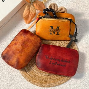 Puede incluir: Una variedad de artículos de cuero, que incluyen una pequeña bolsa naranja con cremallera con "MABON" impreso, un artículo rectangular marrón y un artículo rectangular rojo con el texto "The Lord is my shepherd; I shall not want."