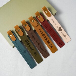 Puede incluir: Cinco marcapáginas rectangulares de cuero con botones a presión de color latón en la parte superior. Los marcapáginas son azul, verde oliva, marrón, rojo oscuro y beige claro, cada uno con texto, fechas, nombres e iconos grabados a láser.