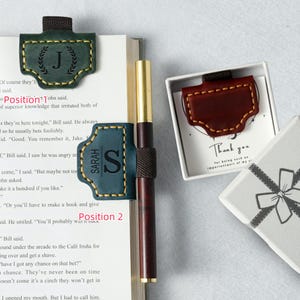 Puede incluir: Una variedad de accesorios de libros de cuero, que incluyen un marcapáginas verde oscuro con la letra "J", un marcapáginas azul con el nombre "SARAH" y un marcapáginas marrón. Un bolígrafo está sujeto por un portalápices de cuero. También hay una caja de regalo.