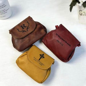 Puede incluir: Tres bolsas de cuero en marrón, burdeos y amarillo. La bolsa marrón tiene una "R" con cuernos. La bolsa burdeos tiene el texto "For God so loved the world... John 3:16". La bolsa amarilla tiene una cruz y el texto "First Confirmation June 11, 2026".