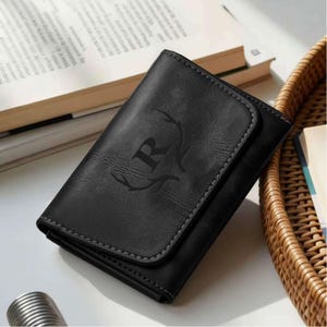 Puede incluir: Cartera de cuero negro con cierre de solapa y costuras grises. La cartera presenta un diseño en relieve de la letra "R" con cuernos de ciervo. La cartera está cerrada y descansa sobre una superficie blanca.
