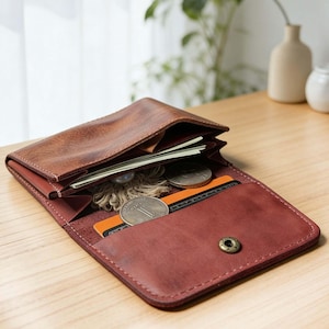 Puede incluir: Cartera de cuero marrón abierta con cierre de botón a presión, que contiene efectivo, monedas y una tarjeta de crédito. La cartera está sobre una superficie de madera, mostrando sus compartimentos interiores y diseño.