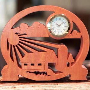 May include: A wooden clock with a farm scene carved into the frame. The clock face is round with Roman numerals and gold trim. The frame is a reddish-brown color and features a sun, clouds, and a house.
