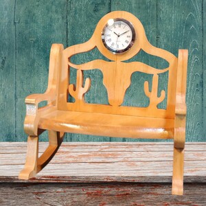 May include: A wooden chair-shaped clock with a light brown finish. The chair back features cut-out designs of cacti and a longhorn skull. A round clock face with Roman numerals is set into the chair's back. The chair is displayed on a wooden surface.