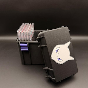 Puede incluir: Caja de almacenamiento negra con un cierre morado, que contiene varias cartas coleccionables. La caja tiene una superficie texturizada y un gráfico blanco y morado de un personaje de dibujos animados en la tapa. La tapa está abierta.