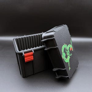 Puede incluir: Caja de almacenamiento negra texturizada con un pestillo rojo y un gráfico verde y negro en la tapa. La caja está abierta, revelando el interior. La caja está diseñada para el almacenamiento.