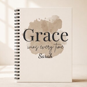 May include: A spiral-bound notebook with a cream-colored cover featuring a beige watercolor splash design. The text reads "Grace wins every time" in a mix of bold serif and script fonts, with the name "Sarah" printed in black script below.