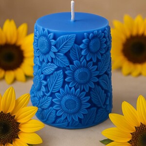 Peut inclure: Une bougie pilier bleue avec un motif de tournesol. La bougie pr&eacute;sente des tournesols et des feuilles en relief. Des tournesols jaunes sont en arri&egrave;re-plan. La bougie a une m&egrave;che blanche.