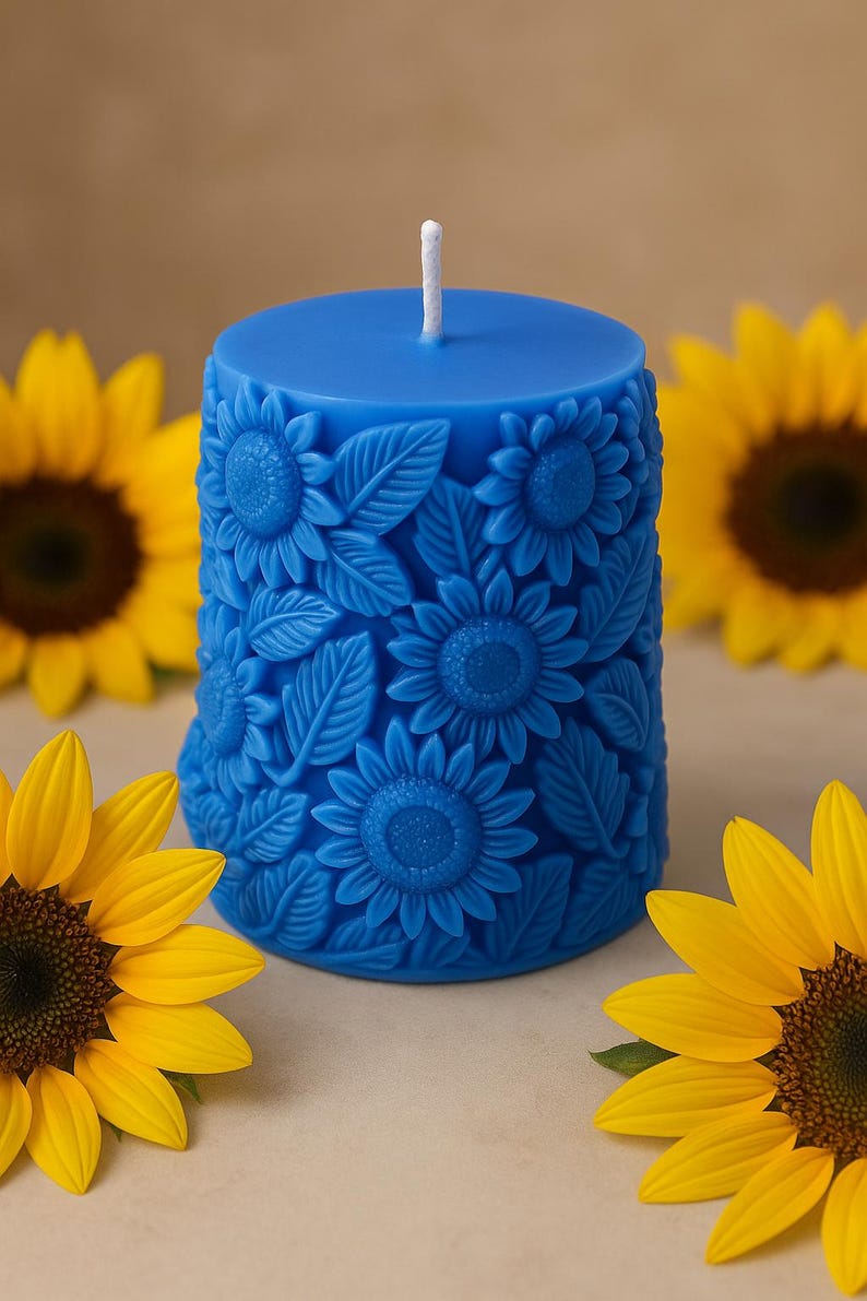 Peut inclure: Une bougie pilier bleue avec un motif de tournesol. La bougie pr&eacute;sente des tournesols et des feuilles en relief. Des tournesols jaunes sont en arri&egrave;re-plan. La bougie a une m&egrave;che blanche.