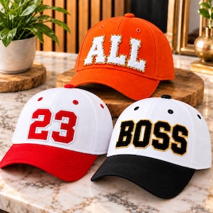 Puede incluir: Tres gorras de béisbol: una naranja con letras blancas que dicen ALL, una blanca con el número 23 en rojo y visera roja, y una blanca con letras negras que dicen BOSS y visera negra. Cada gorra tiene parches bordados.