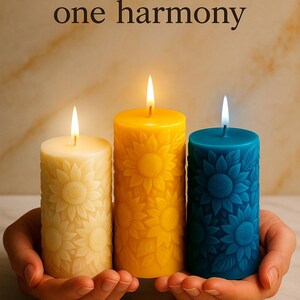 Peut inclure: Trois bougies allum&eacute;es en cr&egrave;me, jaune et bleu sarcelle, chacune avec un motif de tournesol. Les bougies sont tenues dans des mains en coupe. Le texte au-dessus indique : "Trois lumi&egrave;res, une harmonie."