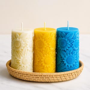 Peut inclure: Trois bougies cylindriques dans un panier tress&eacute;. Les bougies sont cr&egrave;me, jaune et bleu, chacune avec un motif de tournesol. Les bougies ont une m&egrave;che en haut. Le panier est rond et en mat&eacute;riau brun clair. Les bougies mesurent environ 10 cm de haut.