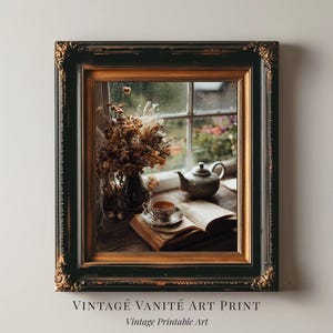 Pode incluir: Uma impressão de arte emoldurada com uma natureza morta com uma xícara de chá, um livro aberto e uma chaleira perto de uma janela. A moldura é preta com detalhes dourados. O texto na parte inferior diz "VINTAGE VANITÉ ART PRINT" e "Vintage Printable Art."