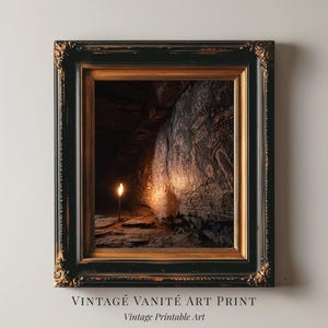 May include: A framed art print featuring a cave interior illuminated by a single candle. The cave wall displays ancient carvings. The frame is black with gold accents. Text at the bottom reads "VINTAGE VANITÉ ART PRINT" and "Vintage Printable Art."