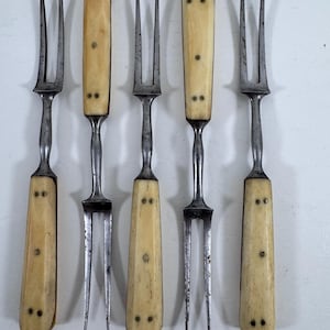 May include: A set of five two-pronged forks with metallic tines and off-white, rectangular handles. Each handle is secured with small metal rivets arranged in a pattern. The metal components show signs of age and wear with a dark, weathered finish.
