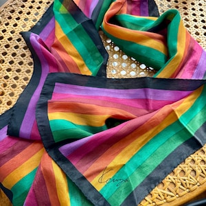 Puede incluir: Una colorida bufanda con rayas de arcoíris en tonos rosa, naranja, verde y morado. La bufanda tiene un borde negro y la firma "Kenzo" en cursiva. La bufanda está sobre una silla de mimbre.
