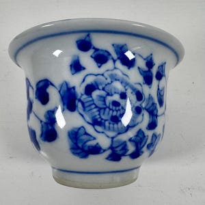 Könnte beinhalten: Kleine, weiße Keramikschale mit blauem Blumenmuster. Die Schale hat eine abgerundete Form mit einem leicht ausgestellten Rand. Das blaue Muster umfasst eine große Blume und Blattranken, was einen klassischen, dekorativen Look ergibt.